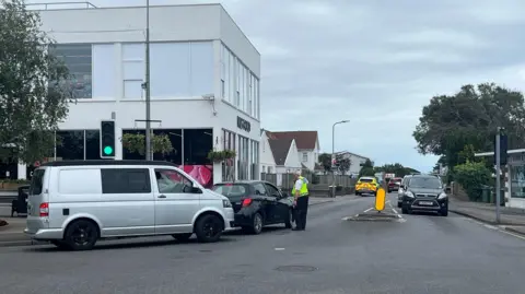 BBC Emergency services are at the scene of a two‑vehicle road traffic collision in Jersey. A police officer is seen directing traffic away from the road closure. 