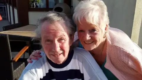 Merseyside Police Eric Greener, who has grey hair swept to the side and is wearing a blue and white striped sweatshirt, smiles at the camera while sitting in a chair outside a pub or restaurant. Sheila Jackson, who has short white hair and is wearing pink cardigan leans into the frame and smiles at the camera with her arm around Greener's shoulder.