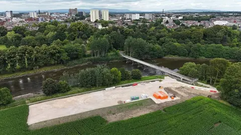 Mike Ashmore Aerial view of the site showing the river with the bridge across it, a large, flat area in the foreground with construction vehicles and metal storage units, and Preston city centre in the background