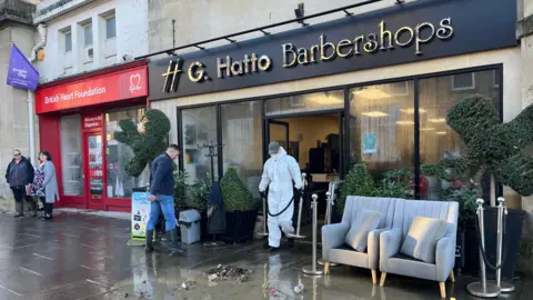 Sofas are out the front of a barbershop and men are walking in and out - one wearing white PPE.