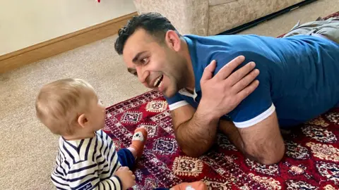 Family handout An Afghan man with short dark hair wearing a blue t-shirt, lying on a rug on the floor, smiling at young child with short fair hair, wearing a white long sleeve t-shirt with blue stripes