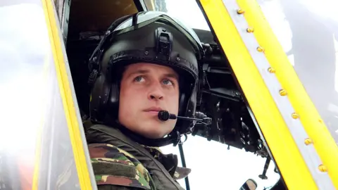 Getty Prince William during in a helicopter during a training exercise. He is wearing a RAF military pattern camouflage uniform and a black helmet with a microphone attched to the side.