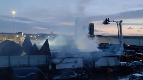 Humberside Fire and Rescue Service A building badly damaged and smoking after a fire. Firefighters can be seen in a raised platform spraying water onto the structure. Burned debris and several parked vehicles surround the scene.