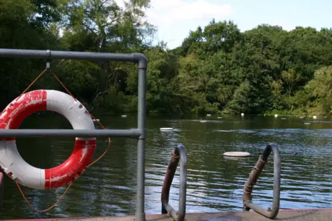 Life ring on a metal railing beside a leafy outdoor bathing pond with steps leading into the water at Hampstead Heath