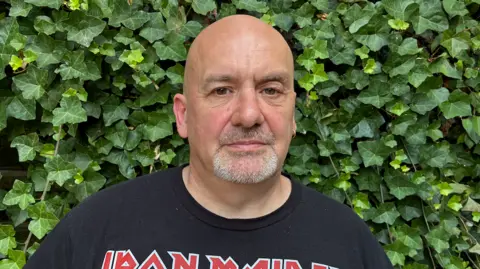 Cathy Killick/BBC A white man with a short grey beard and moustache, and an Iron Maiden t shirt, looking at the camera. 