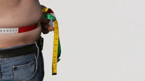 A man holding a tape measure around his bare tummy. He is wearing jeans.