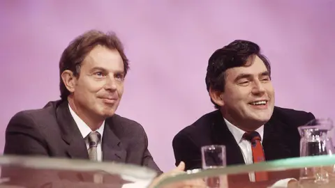 Tony Blair is sat connected  the near  of Gordon Brown. Blair is wearing a grey suit   and Brown is wearing a achromatic  suit. The brace  are smiling and looking retired  towards an audience