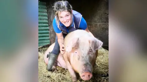 Ellie Maguire Ellie Maguire is wearing wellies, trousers and a blue shirt under a gilet. She is smiling whilst her arms are around a large pig.