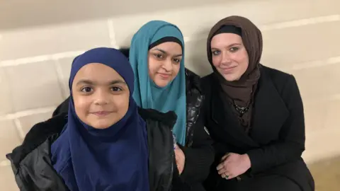 Nisha wearing a blue scarf, Maira, a young girl in a blue scarf and black coat and Eva, wearing a brown scarf, smiling for the camera.