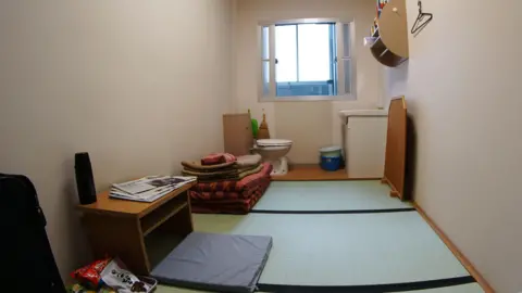 Getty Images A spare room with bamboo mat on the floor, toilet and sink at the back, neatly piled bedding and window onto a corridor beyond