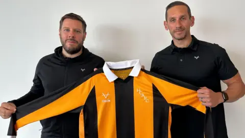 Matt Dean / BBC Lee Jenkinson and another man holds the new Hull City shirt which has black and amber vertical stripes and a white collar. The men have short dark hair and beards and wear black clothes