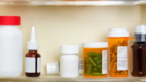 A medicine cabinet shelf with three orange containers of medicine, a bottle with a cap, one with a pipette top and a tube of cream behind them. Some tablets are visible in the transparent orange bottles.