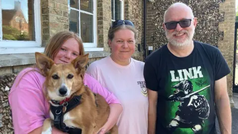 Vikki Irwin/BBC A young woman holding a dog stands beside her mother and father who are all smiling at the camera. The young woman has long red hair and is wearing a pink top. The mother has tied up blonde hair and wears a lighter pink T-shirt with sunglasses on her head. The man is bald with a grey beard, sunglasses and a black T-shirt.