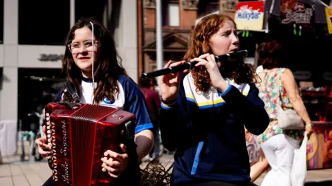 Two young women. One is playing an accordian, the other is playing a flute.
