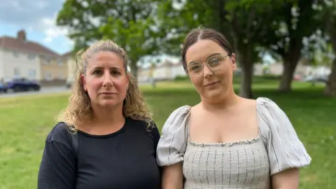 Two women in front of the camera, the one on the left wears a black top and has curly blonde hair. The woman on the right wears a grey dress, she wears glasses and has dark hair. In the background there are trees and greenery.