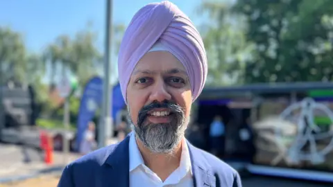 Slough MP Tan Dhesi is wearing a pink turban as well as a white shirt and navy suit. He is smiling and the pop-up heart clinic is visible in the background. 