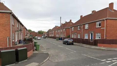 Google Morris Street in Gateshead. The picture is taken from the junction of the road and shows semi-detached housing lining the street. Several cars are parked outside and green wheelie bins have been put out for collection.