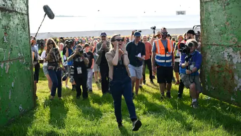 Emily Eavis, co-organiser of the Glastonbury Festival, officially opens the gates at Worthy Farm for the festival, in Pilton, Somerset. She is standing at the front of a crowd of photographers and shouting with her hands around her mouth, while kicking a leg out.