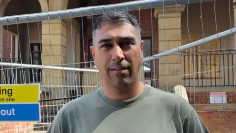 A man in a green t-shirt stands in front of railings next to a large old brick building 