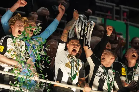 Owen Humphreys / PA Media Newcastle hold the Carabao Cup aloft in victory. Team members Anthony Gordon, Nick Pope, Bruno Guimaraes, Kieran Trippier and Dan Burn are standing on the a balcony, looking on at cheering fans. They are wearing black and white, with Pope is wearing the blue goalkeeper kit, while green and white confetti shoots up in front of them. 