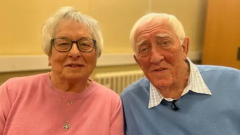Wendy, wearing a pink top, and Ken, in a blue jumper, are sitting in a police station looking at the camera during their interview.