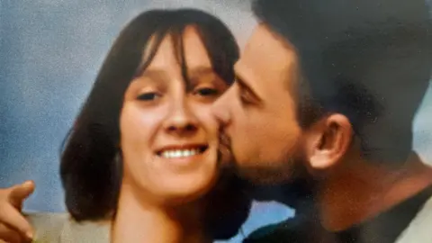 Family photo A woman with a brunette bob haircut is smiling at the camera. She is being kissed on the cheek by a younger man who has dark hair and a beard.