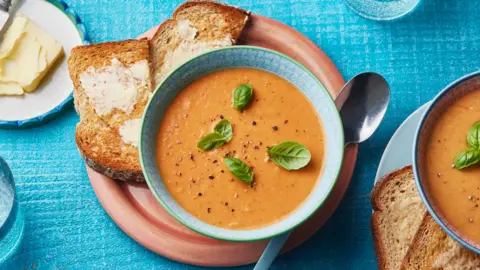 A top-down photo showing a bowl of tomato soup on a plate with toast and a spoon.