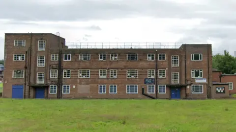 Google A red-brick building with three storeys and a four-storey tower at one end. It has rows of white-framed windows with one boarded up.