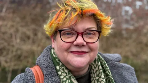 BBC/Paul O'Gorman Annie Wynn, smiling wearing glasses. She has bright yellow hair with orange streaks in it, and is wearing a grey coat and green scarf.