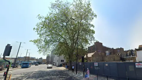 Just Giving A large tree with lots of green leaves on a pavement. In the photo there are houses and a traffic light junction. There is a woman waiting at the bus stop.