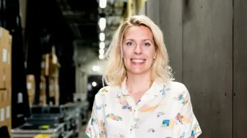 Sara Pascoe smiles in a backstage area - she has blonde hair and wears a white shirt with multicoloured patterns