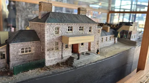 A model railway station sits inside a wood and glass display case. The model biggest in shot is of the station building a grey brick structure with a tiled roof. 