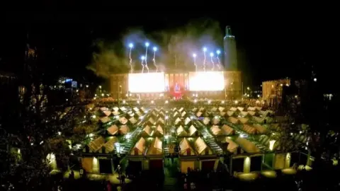 An aerial view of Norwich market. The stall roofs are visible and are lit up by an explosion of light and fireworks 