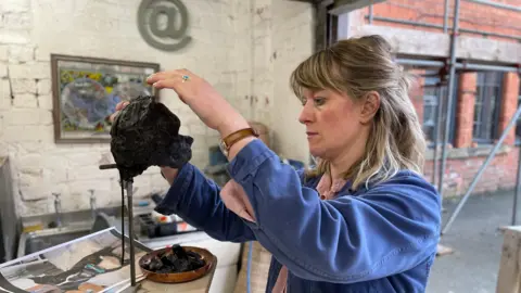 Mark Ansell / BBC Sculptor Lily Marsh, wearing a pink shirt and blue overcoat, models a black sculpture of a dog's head in her studio.