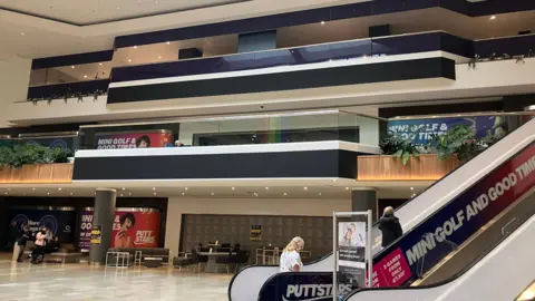 The exterior, inside the shopping centre, of the former John Lewis store with grey shutters down over its main entrance. In the foreground is an escalator with some people going up it.
