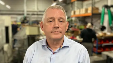 Sam Read/BBC A man wearing a light blue shirt looking into the camera. He has short grey hair and is standing in a factory setting with work going on behind him.