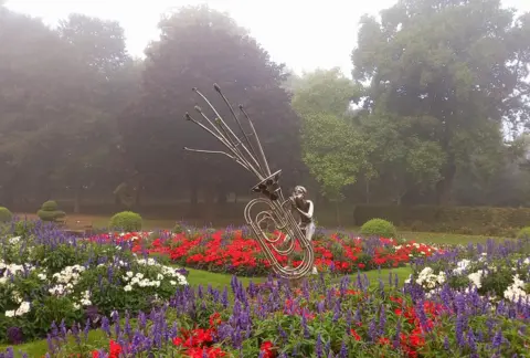 BBC Weather Watchers/Banjo calling A metal sculpture of a child playing a tuba, standing in a park with beds of red, white and blue flowers around it.