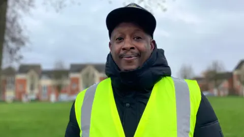 Carlos Dyer, in warm clothing and wearing a high-vis vest, with a park area in the background, looking at the camera