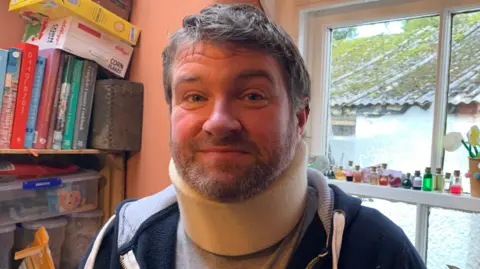 Dafydd Morris Jones smiling looking at the camera. He is stood in front of his window. On the left, behind him, there is a book shelf with various books and toys on. Dafydd is wearing a grey t-shirt, a navy zipper hoodie, and a cream neck brace. 