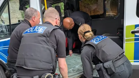 Four people in black police uniforms lifting a memorial plaque out of the side of a police van. The plaque is large and appears blue with age.