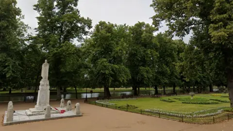 A war memorial, which is of a man and is made of grey stone, is on the left. Behind the statue is a river which goes in between a field one side and a walk way the other. 