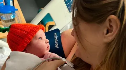 Family photo Natalie looking down at newborn baby Arthur in her arms wearing an orange hat. She has brown hair and a blood pressure monitor on her finger and is sat in a hospital bed, with a wooden table and chair beside her.