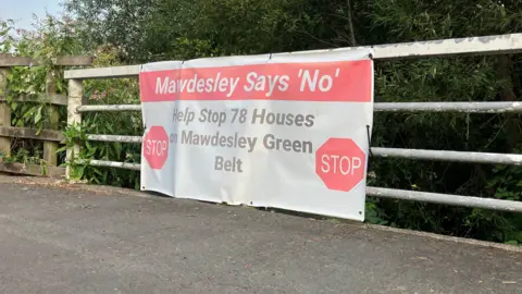 A banner on a metal fence, that says "Mawdesley says no - help stop 78 houses on Mawdesley Green Belt" with two stop signs as decoration.