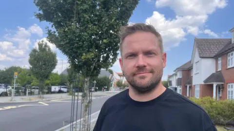 A young man in a navy T-shirt looking at the camera with slight smile. He stands on a sunny tree-lined street surrounded by new homes