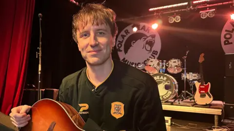 A man with short light brown hair and wearing a black Hull City shirt with an orange badge is standing in front of a stage and holding a guitar. There is a black backdrop with a drum kit and a guitar, as well as a microphone stand.