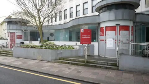 The front of Philip Le Feuvre House. The building has a grey ground level facade with two steps leading to the main entrance. The upper part of the building sits slighty behind the entrance and is white stone with black-framed windows. There is red signage in front of the entrance with the words 'Customer and Local Services'.