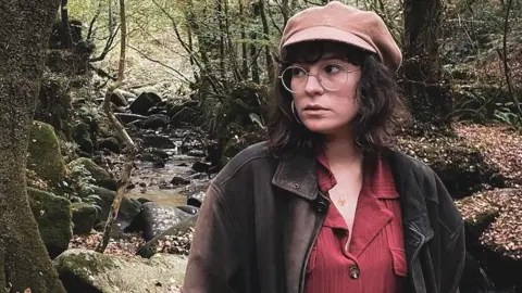 ALICE DUDLEY Alice, a woman with shoulder-length brown hair, is wearing a beige cap and large glasses. She looks to one side and is standing in a glen, with a shallow stream behind her.