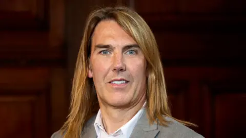 Wakefield Council A man with long golden hair looking into the camera wearing a grey suit and white shirt.