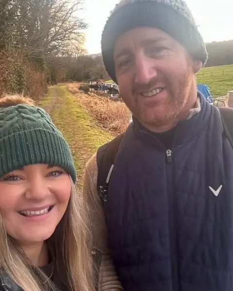Siobhan Gorman A selfie of Siobhan and her partner who are in the countryside with a canal behind them. Both are smiling at the camera and wearing bobble hats. She has long wavy blonde hair and is smiling. He has a cropped beard.