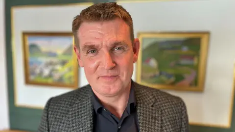 Faroese prime minister Aksel Johannesen pictured in an office with two blurred out gold-framed paintings hanging on a white wall behind him. He has brown hair combed to the side and is clean-shaven. He is looking directly at the camera with a serious expression. He is wearing a black shirt and a checked blazer.